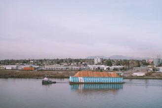 A small tugboat is guiding a large barge laden with what appears to be a pile of earth or sawdust. The barge is labeled with 'Ledcor Group' on its side. In the background, a cityscape is visible with numerous buildings, industrial structures, and a wooded area. Mountains rise in the distance beneath a cloudy sky.