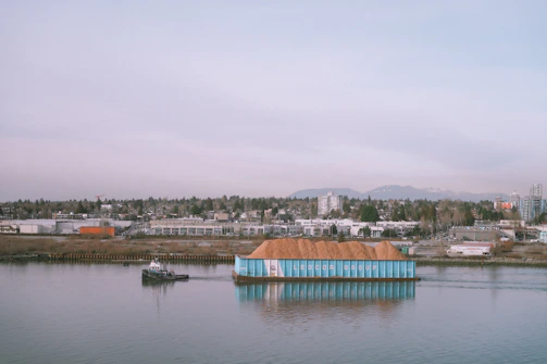 A small tugboat is guiding a large barge laden with what appears to be a pile of earth or sawdust. The barge is labeled with 'Ledcor Group' on its side. In the background, a cityscape is visible with numerous buildings, industrial structures, and a wooded area. Mountains rise in the distance beneath a cloudy sky.