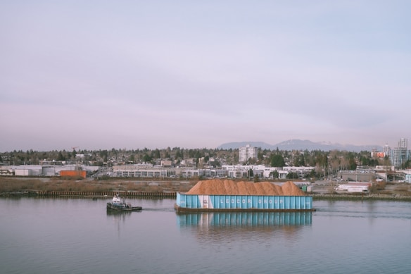 A small tugboat is guiding a large barge laden with what appears to be a pile of earth or sawdust. The barge is labeled with 'Ledcor Group' on its side. In the background, a cityscape is visible with numerous buildings, industrial structures, and a wooded area. Mountains rise in the distance beneath a cloudy sky.
