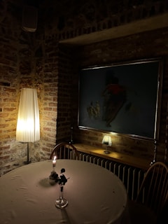 A dimly lit room with exposed brick walls, featuring a round table covered with a cloth and a small vase holding a single flower under candlelight. A tall floor lamp provides warm lighting beside the table. On the wall, a painting is partially illuminated. A smaller decorative lamp sits on a shelf next to the painting, adding to the cozy ambiance.