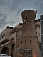 A large windmill structure is integrated into a building that houses a bakery. The windmill's blades are illuminated, contrasting against the dusk sky. The structure features brickwork and modern design elements, with signage for 'Wooden Bakery' visible near the entrance. The sky is cloudy, adding to the evening ambiance.