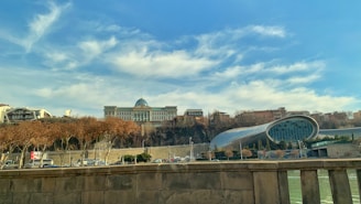 A large, classical-style building with a dome is positioned on a hill surrounded by trees. In the foreground, there's a modern, cylindrical glass structure, likely a theater or concert hall. Trees with autumn leaves line the road below, and a stone railing is visible, suggesting a bridge or elevated walkway.