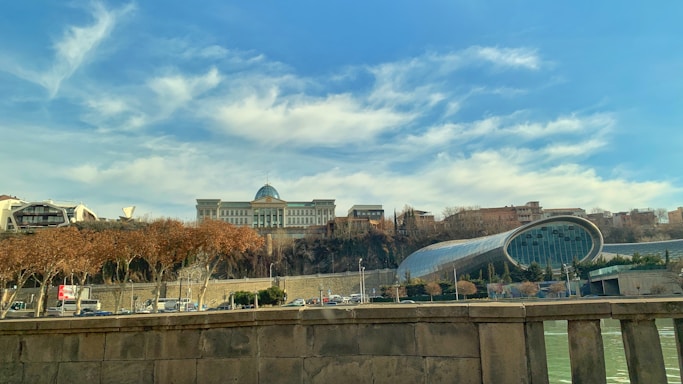 A large, classical-style building with a dome is positioned on a hill surrounded by trees. In the foreground, there's a modern, cylindrical glass structure, likely a theater or concert hall. Trees with autumn leaves line the road below, and a stone railing is visible, suggesting a bridge or elevated walkway.