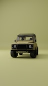 A vintage-style off-road vehicle is prominently displayed from the front on a minimalistic, uniform olive green background. The vehicle is tan with a sturdy frame, circular headlights, and rugged tires, creating a sense of adventure and durability.