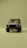A vintage-style off-road vehicle is prominently displayed from the front on a minimalistic, uniform olive green background. The vehicle is tan with a sturdy frame, circular headlights, and rugged tires, creating a sense of adventure and durability.