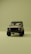 A vintage-style off-road vehicle is prominently displayed from the front on a minimalistic, uniform olive green background. The vehicle is tan with a sturdy frame, circular headlights, and rugged tires, creating a sense of adventure and durability.