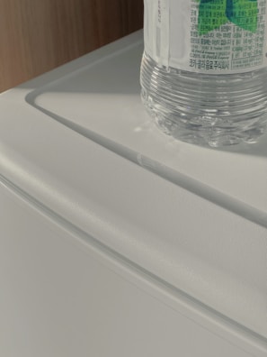 A clear plastic bottle with a label featuring Korean text rests on a white, textured surface. The scene is illuminated by natural sunlight, casting soft shadows.