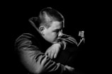 A black and white portrait of a person with short hair resting their head on their arm while holding a guitar. The person is wearing a textured jacket and appears thoughtful or contemplative.