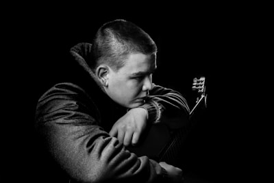 A black and white portrait of a person with short hair resting their head on their arm while holding a guitar. The person is wearing a textured jacket and appears thoughtful or contemplative.