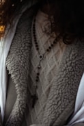 A close-up view of a person wearing a textured knit sweater with a rosary featuring wooden beads and a cross pendant draped over it. The outerwear appears to have a wool or fur-lined interior.