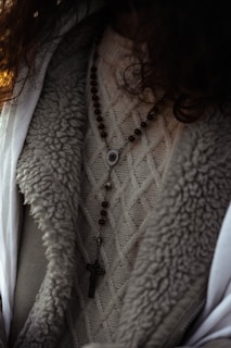 A close-up view of a person wearing a textured knit sweater with a rosary featuring wooden beads and a cross pendant draped over it. The outerwear appears to have a wool or fur-lined interior.