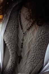 A close-up view of a person wearing a textured knit sweater with a rosary featuring wooden beads and a cross pendant draped over it. The outerwear appears to have a wool or fur-lined interior.