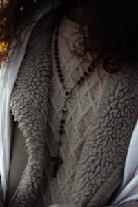 A close-up view of a person wearing a textured knit sweater with a rosary featuring wooden beads and a cross pendant draped over it. The outerwear appears to have a wool or fur-lined interior.