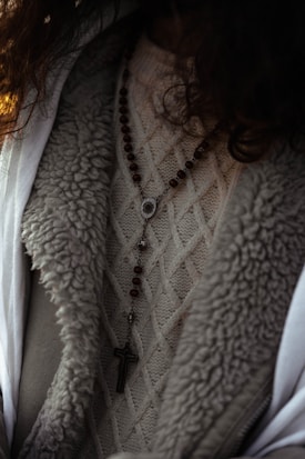 A close-up view of a person wearing a textured knit sweater with a rosary featuring wooden beads and a cross pendant draped over it. The outerwear appears to have a wool or fur-lined interior.