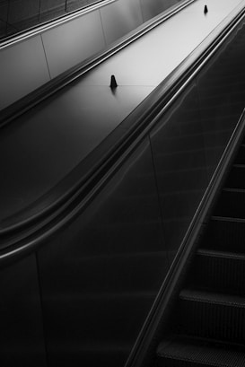 Black and white image of an escalator with clean, smooth lines and a modern design. The perspective focuses on the handrails and the stairs, leading the eye upwards.