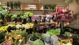 An indoor display features a variety of artificial flowers and plants arranged on wooden shelves. The shelves are filled with vibrant green, white, purple, and red flowers. There are also several decorative items, including golden elephant figurines and Buddha head statues, placed among the greenery.