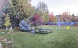 A serene backyard garden featuring a variety of trees and shrubs, including evergreens and deciduous trees with autumn leaves. The grass is well-maintained, and a small round table with two bright yellow chairs is set up on the lawn. A decorative statue is placed among some plants, and there's an umbrella stand without the canopy.