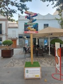 A multi-directional signpost in a public square with various words such as 'Empatía,' 'Diversidad,' and 'Equidad,' each on different colored arrows. The signpost is surrounded by outdoor seating, with tables and chairs set up under a large umbrella. The area is paved with decorative stones and flanked by buildings, one of which features a balcony. Leafy trees partially frame the scene, adding a touch of greenery.