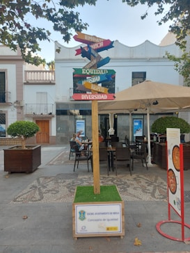 A multi-directional signpost in a public square with various words such as 'Empat&iacute;a,' 'Diversidad,' and 'Equidad,' each on different colored arrows. The signpost is surrounded by outdoor seating, with tables and chairs set up under a large umbrella. The area is paved with decorative stones and flanked by buildings, one of which features a balcony. Leafy trees partially frame the scene, adding a touch of greenery.