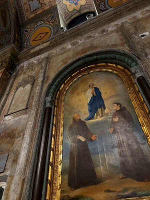 A religious painting on an ornate wall depicts a central figure in blue robes surrounded by cherubs, poised above two figures dressed in monastic robes, part of a grand architectural setting with decorative arches and symbols.