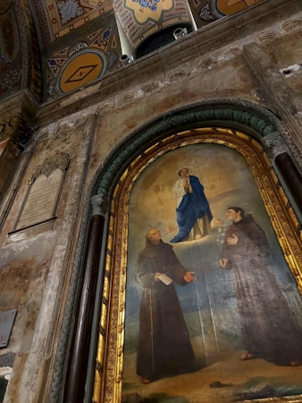 A religious painting on an ornate wall depicts a central figure in blue robes surrounded by cherubs, poised above two figures dressed in monastic robes, part of a grand architectural setting with decorative arches and symbols.