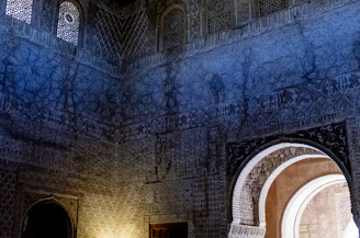 The image depicts an intricately designed interior wall featuring detailed geometric and floral patterns. The wall is illuminated by natural light filtering through ornate windows, casting shadows on the surface. An arched doorway leads to a sunlit area beyond, adding contrast to the darker interior.