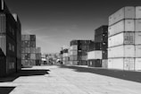 Stacks of shipping containers are arranged in rows, creating an industrial atmosphere. A forklift is visible in the distance among the containers, on a concrete surface under a clear sky.