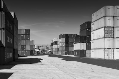 Stacks of shipping containers are arranged in rows, creating an industrial atmosphere. A forklift is visible in the distance among the containers, on a concrete surface under a clear sky.