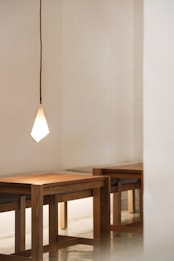 A minimalist room with light-colored walls features a wooden table and chairs. A geometric-shaped pendant light hangs from the ceiling, casting a soft glow. The floor appears to be polished, reflecting some light.