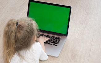 A young child with blonde hair tied in a ponytail is sitting on the floor, using a laptop with a bright green screen. The child is wearing a white shirt and their attention is focused on the laptop. The floor appears to be a light wooden texture, creating a neutral background for the scene.