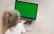 A young child with blonde hair tied in a ponytail is sitting on the floor, using a laptop with a bright green screen. The child is wearing a white shirt and their attention is focused on the laptop. The floor appears to be a light wooden texture, creating a neutral background for the scene.