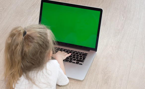 A young child with blonde hair tied in a ponytail is sitting on the floor, using a laptop with a bright green screen. The child is wearing a white shirt and their attention is focused on the laptop. The floor appears to be a light wooden texture, creating a neutral background for the scene.