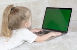 A young child with long blonde hair is lying on a light-colored sofa while using a laptop. The laptop screen is bright green, suggesting it might be a green screen or placeholder for an image.