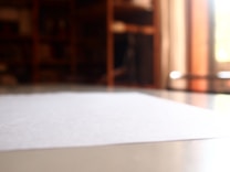 A close-up view of a blank white paper on a reflective surface, with a blurred background that includes shelves and warm lighting.