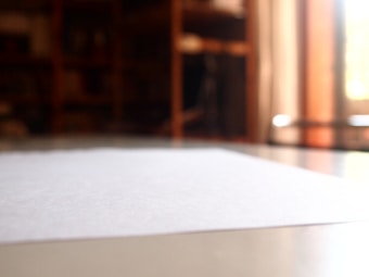 A close-up view of a blank white paper on a reflective surface, with a blurred background that includes shelves and warm lighting.
