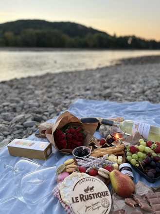 A picturesque picnic setup on a rocky riverside at sunset. The spread includes red roses, a variety of cheeses, figs, grapes, pears, strawberries, macarons, chocolate, and a bottle of white wine. Also present are a wooden serving board, a candle, and two wine glasses.