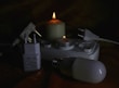 Several electrical items including plugs and a power strip are arranged next to two lit candles of different sizes. The items include a white plug adapter, a power strip with multiple sockets, and a light bulb. The setup appears to be in a dimly lit environment with the candles providing the main source of light.