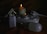 Several electrical items including plugs and a power strip are arranged next to two lit candles of different sizes. The items include a white plug adapter, a power strip with multiple sockets, and a light bulb. The setup appears to be in a dimly lit environment with the candles providing the main source of light.