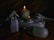 Several electrical items including plugs and a power strip are arranged next to two lit candles of different sizes. The items include a white plug adapter, a power strip with multiple sockets, and a light bulb. The setup appears to be in a dimly lit environment with the candles providing the main source of light.