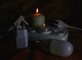 Several electrical items including plugs and a power strip are arranged next to two lit candles of different sizes. The items include a white plug adapter, a power strip with multiple sockets, and a light bulb. The setup appears to be in a dimly lit environment with the candles providing the main source of light.