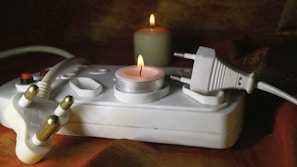A white power strip with multiple electrical outlets is placed on a surface. A lit candle is positioned on the power strip, casting a warm glow. Another candle is burning in the background, adding to the low-light, cozy ambiance. Two unconnected plugs are visible, one partially inserted into the power strip.