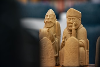 Two intricately carved chess pieces with detailed facial expressions stand prominently. The pieces appear to be made of a light-colored material, possibly wood or ivory, and feature a bishop and a warrior figure. Their expressions are serious and attentive, indicative of historical or strategic significance.
