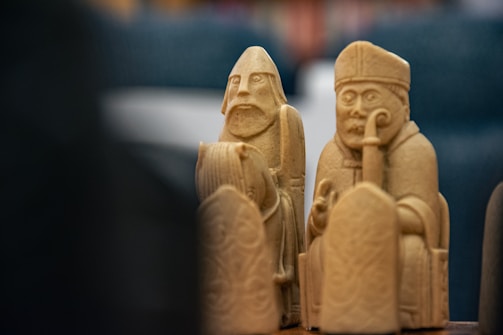 Two intricately carved chess pieces with detailed facial expressions stand prominently. The pieces appear to be made of a light-colored material, possibly wood or ivory, and feature a bishop and a warrior figure. Their expressions are serious and attentive, indicative of historical or strategic significance.