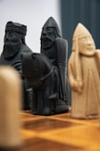Intricately carved chess pieces resembling medieval warriors are set on a wooden chessboard. The focus is on the detailed features of the figures, which include carved facial expressions and armor. The background is slightly blurred, highlighting the craftsmanship of the pieces.