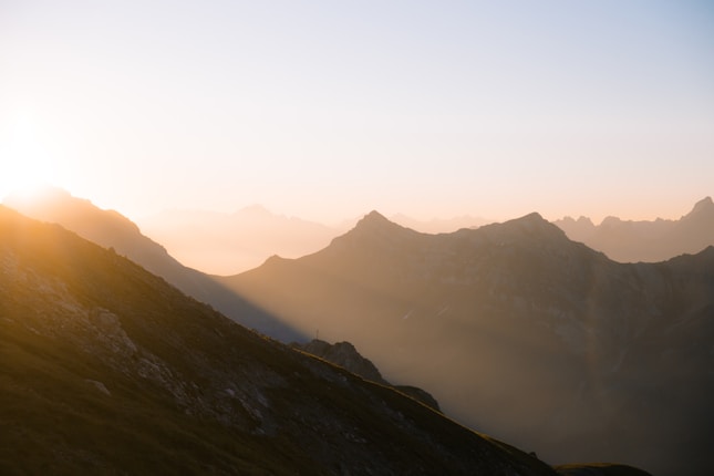 A serene mountain landscape at sunrise with sunlight casting a warm glow on the peaks. The sky transitions from soft pinks and oranges to a clear blue as the sun emerges from behind the mountains.