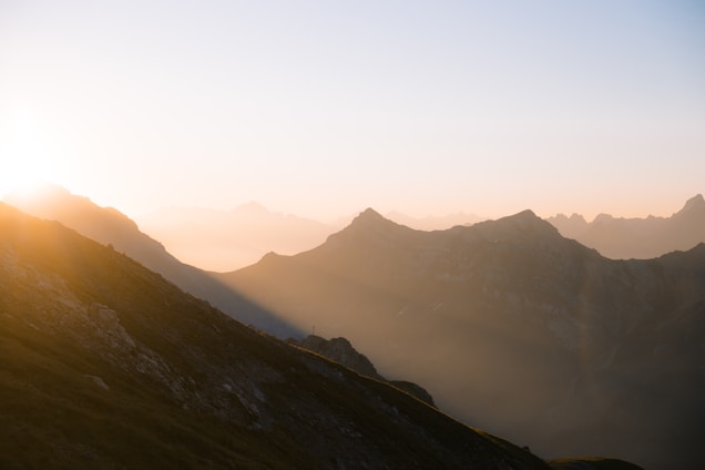 A serene mountain landscape at sunrise with sunlight casting a warm glow on the peaks. The sky transitions from soft pinks and oranges to a clear blue as the sun emerges from behind the mountains.