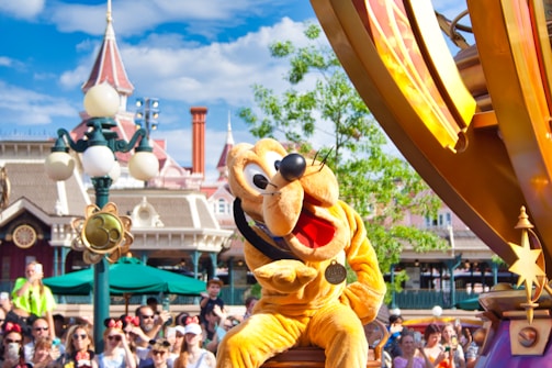 A vibrant amusement park setting featuring a large cartoon character statue of a dog wearing a name tag displayed prominently in the foreground. The scene is lively with a crowd of people in the background, many wearing themed clothing and accessories. Behind the character, a whimsical structure with turrets and a lamppost adds to the festive atmosphere, and the sky is clear with fluffy clouds.