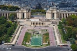 An aerial view of an urban park with expansive green lawns, pathways, and fountains surrounded by historic architecture. Numerous people are seen walking along the paths, and the cityscape stretches into the background filled with trees and buildings.