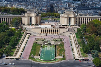 An aerial view of an urban park with expansive green lawns, pathways, and fountains surrounded by historic architecture. Numerous people are seen walking along the paths, and the cityscape stretches into the background filled with trees and buildings.