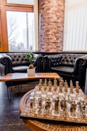 A luxurious interior features two black leather tufted sofas arranged around a wooden coffee table, topped with a small potted plant. In the foreground, an ornate chess set with metal pieces is displayed on a patterned board. The background includes a large window with a view of a cityscape and a prominent brick column.